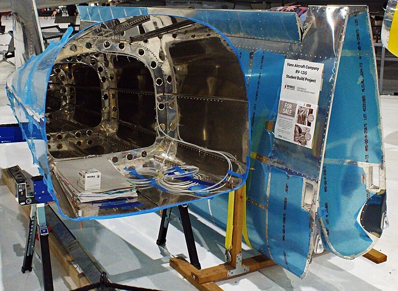 A Van's RV-12iS fuselage built by students at the Wings Over the Rockies Air and Space Museum, Denver, June 2025: rows of rivets along the skins and bulkheads, the protective blue film still on the aluminum, and a wire bundle laid in for routing. Photo: ZLEA, CC BY-SA 4.0, via Wikimedia Commons (cropped).